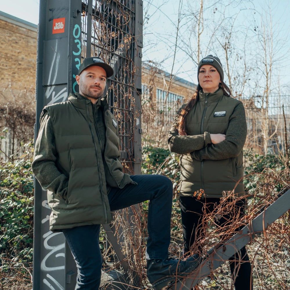Zwei Personen in modischer Outdoor-Bekleidung stehen an einem graffitibedeckten Stahlgerüst im Freien.